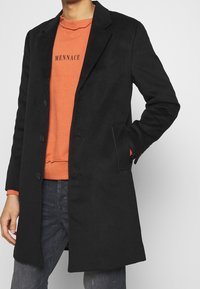 Černý kabát z vlněné směsi s výrazným designem, klopami, třemi knoflíky, bočními kapsami a přiléhavým střihem přes oranžovou mikinu.