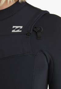 Nahaufnahme eines schwarzen Neoprenanzugs-Schulters mit Wellen-Logo und geschlungener Riemen-Detail an der Schulternaht.