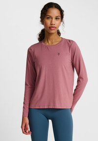 Kvinnors långärmad topp i ljusrosa tyg, med rund halsringning och logodetalj på bröstet, bärs med teal leggings.
