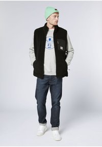 Black fleece vest with full zip, side pockets, and green nylon patch, worn over a grey sweatshirt with blue graphic design and blue jeans.