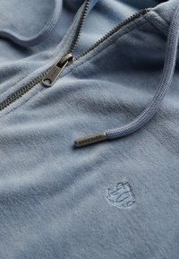 Sudadera con cremallera de color azul claro hecha de una tela suave, con un pequeño logo bordado y una cremallera metálica plateada con un cordón texturizado.