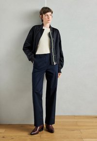 Geacă bomber din satin negru peste un top crem cu dungi, asortată cu pantaloni largi de culoare bleumarin și cizme cu toc închis burgundy. Fundal cu podea din lemn.