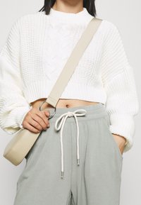 Weißer, kurz geschnittener Pullover mit gerippter Textur und Zopfstrickdetails. Kombiniert mit hellgrauen Hosen und einer kleinen, beigefarbenen Umhängetasche.