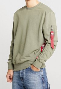 Sudadera verde oliva hecha de una mezcla de algodón, con un bolsillo con cremallera en la manga y un acento lateral rojo con texto. Puños y dobladillo acanalados.