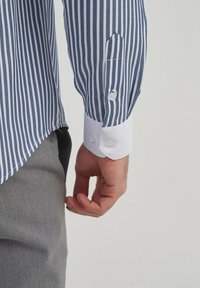 Chemise rayée bleue et blanche avec une coupe ajustée, dotée d'un poignet blanc et d'une fermeture à boutons, avec une texture de tissu lisse.