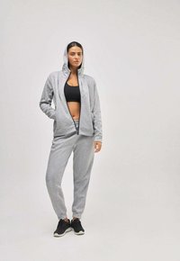 Sudadera gris con cremallera y capucha, combinada con pantalones de chándal a juego. Una camiseta de deporte negra y zapatillas negras completan el conjunto. Tejido suave y texturizado.
