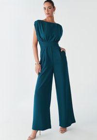 Model wearing a teal jumpsuit with a fitted waist and wide legs, standing confidently against a light background, accented by gold jewelry.