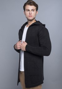 Sort strikket cardigan med hætte, med ribbede manchetter og åben front, båret over en hvid T-shirt, set fra siden.