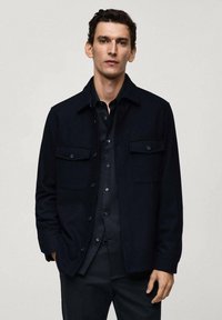 Donkerblauwe jas van structuurstof, met een knoopsluiting, twee borstzakken en een kraaghals. Gestyled met een zwart shirt.