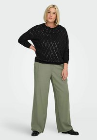 Črn pulover z diamantskim vzorcem, ohlapne prileganje in dolgimi rokavi; kombiniran s svetlo olivno širokimi hlačami in črnimi čevlji.