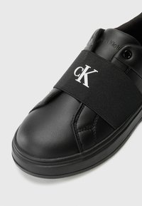 Baskets noires avec tige en cuir et bande élastique, arborant le logo "cK" en blanc. Bout arrondi avec surface texturée et semelle en caoutchouc noire.