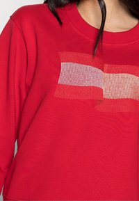 Rode sweatshirt met ronde halslijn en een textuurontwerp van een vlag in lichtrood en crème over de voorkant. Zachte stof, ontspannen pasvorm.