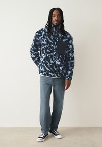 Pulloverjack in marineblauw en lichtblauw tie-dye, gemaakt van fleece, met een ritskraag en een borstzak. Draagt lichtblauwe jeans en zwarte sneakers.