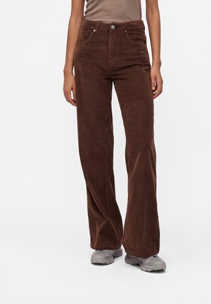 Personne portant un pantalon en velours côtelé marron taille haute et des baskets grises, debout devant un fond blanc uni.