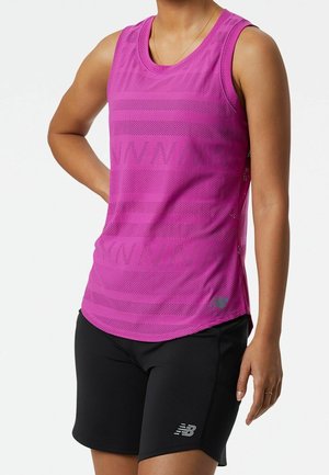 Vrouw draagt een mouwloze roze geperforeerde sporttop en zwarte New Balance short, staand met één hand op haar heup.