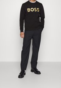 Person som bär en svart sweatshirt med "BOSS" i gyllene bokstäver, ihop med mörka byxor och glänsande svarta skor, stående mot en enkel bakgrund.