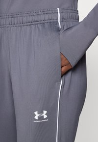 Šedé športové nohavice s úzkym strihom, elastickým pásom, bočnou kapsou, bielymi akcentmi a logom Under Armour na dolnej časti nohavice.