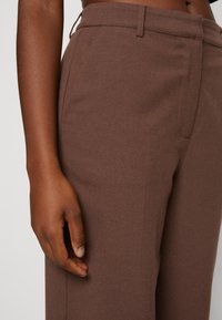 Pantalones marrones a medida con una superficie lisa y texturizada, que presentan un delantero plano y bolsillos laterales, acompañados de una mano descansando sobre la tela.