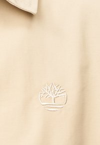 Tissu beige avec un logo circulaire blanc cousu, présentant un arbre stylisé avec des branches et des lignes ondulées en dessous.