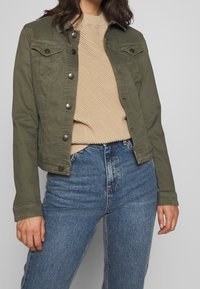 Oliven grøn denimjakke med trykknapper, to brystlommer og et cropped design, parret med en beige tekstureret trøje og blå højalpine jeans.