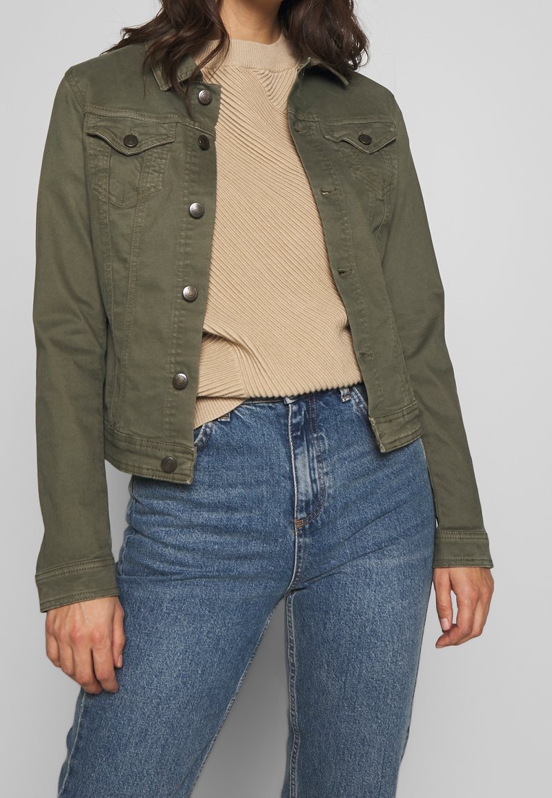 Oliven grøn denimjakke med trykknapper, to brystlommer og et cropped design, parret med en beige tekstureret trøje og blå højalpine jeans.