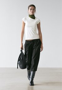 Femme marchant, portant un haut blanc à manches courtes, un pantalon noir coupé, des bottes noires, une écharpe verte et un grand sac à main noir.