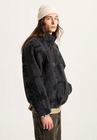 Schwarze Fleecejacke mit einer strukturierten, plüschigen Außenseite und einem einfarbigen schwarzen Einsatz auf der Brust. Das Model trägt eine hellfarbige Strickmütze.