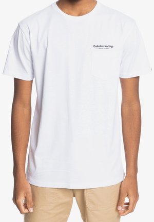 Młoda osoba nosząca gładki biały t-shirt z krótkim rękawem z małym logo Quiksilver 1969 nad lewą kieszenią na piersi oraz beżowe spodnie.