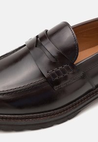Zwart gepolijst leren penny loafer met gestikte details, ronde neus en dikke rubberen zool op een witte achtergrond.