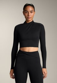 Fekete, hosszú ujjú, rövidített topp, magas gallérral, ahol a tervezés egy átkötött stílust mutat be, sima textúrával, amelyhez magas derekú leggings tartozik, csavart részlettel.