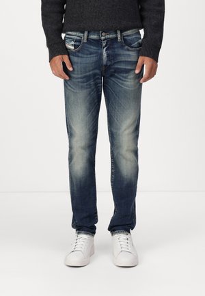 Personne portant un jean bleu délavé avec des baskets blanches et un pull sombre, debout devant un fond clair uni.