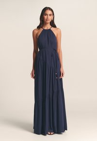 Mörk marinblå maxiklänning med halterneck, rynkat tyg och diagonala paneler. Utrustad med ett knytskärp i midjan och dekorativa gulddetaljer.