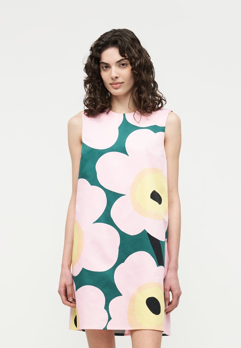 Νεαρή γυναίκα με σγουρά μαλλιά που φοράει αμάνικο, μέχρι το γόνατο φόρεμα με μεγάλο μοτίβο ρόζ και κίτρινων λουλουδιών σε πράσινο φόντο.