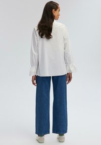 Blusa bianca con dettaglio del colletto ricamato e pizzo sui polsini, abbinata a jeans blu a gamba larga e sneakers bianche.