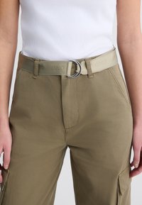 Khakifarbene Cargo-Hosen mit Seitentaschen, kombiniert mit einem hellbeigen Gürtel mit einer runden silbernen Schnalle und geprägtem Logo.