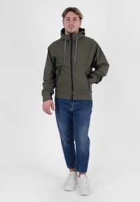 Olivgrüne Kapuzenjacke mit einem Frontreißverschluss, zwei Seitentaschen und grauen Kordeln, kombiniert mit blauen Jeans und weißen Turnschuhen.