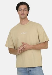 Beige Baumwoll-T-Shirt mit kurzen Ärmeln, lockerer Passform, runder Ausschnitt und weißem Text-Detaillierung auf der Brust. Getragen mit hellblauen Denim-Jeans.