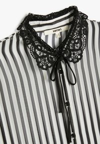 Blusa a rayas en blanco y negro con un cuello de encaje negro, detalle de lazo y botones negros. El material parece ligero y transparente.