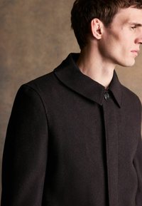 Czarny wełniany płaszcz z wysokim kołnierzem, zapięciem na guziki i gładką fakturą. Dopasowany krój, który podkreśla minimalistyczny design z czystymi liniami.