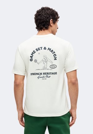 Homme portant un t-shirt blanc avec une illustration d'un joueur de tennis et le texte "Game Set & Match, French Heritage Lacoste Club" dans le dos.