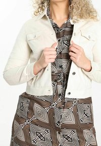 Creme denimjakke med to brystlommer, knaplukning og lange ærmer, lagd over en mønstrets brun kjole med sorte geometriske former.