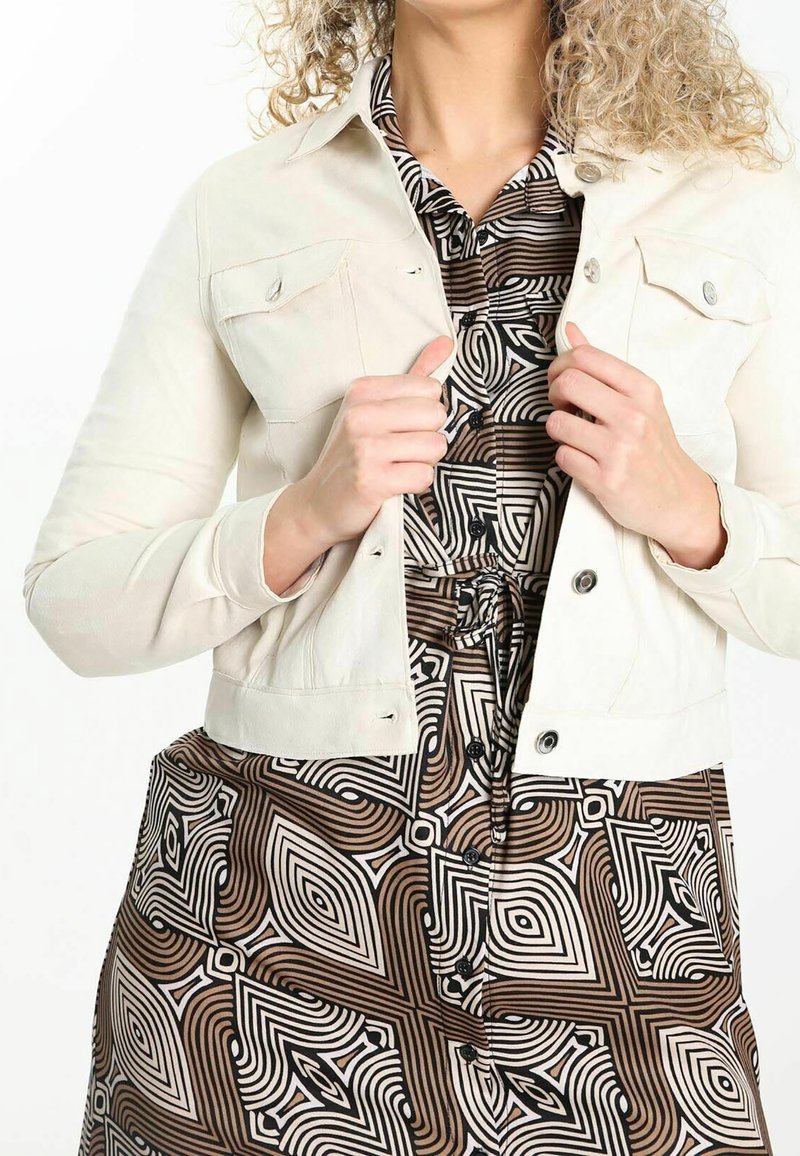 Creme denimjakke med to brystlommer, knaplukning og lange ærmer, lagd over en mønstrets brun kjole med sorte geometriske former.