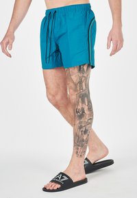 Pantaloni da bagno blu con cintura regolabile, caratterizzati da strisce laterali nere. Abbinati a ciabatte nere. Tatuaggio dettagliato sulla gamba visibile.