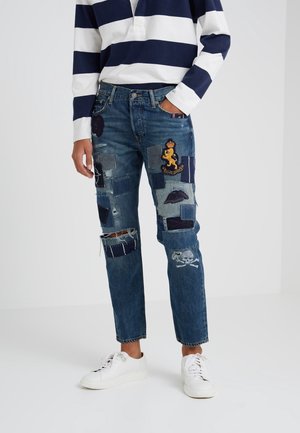 Personne portant un jean patchwork bleu orné de motifs brodés de lion et de crâne, assorti à des baskets blanches et un T-shirt rayé bleu marine et blanc.