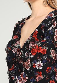 Mustat kukalliset mekko punaisilla ja valkoisilla kukilla. Siinä on rypytetyt reunat ja pitsikoristeet kaula-aukon ympärillä. Läheltä otettu kuva suunnittelusta.