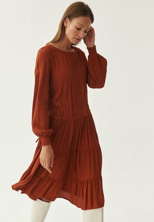Rust-colored dress with long, gathered sleeves, a round neckline, and tiered ruffles at the hem. Soft, lightweight fabric with a smooth texture.