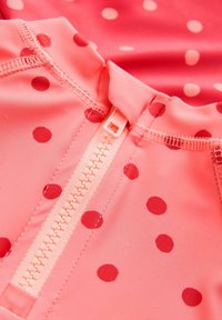 Tissu rose à pois avec une fermeture à glissière à l'avant. Le matériau est lisse avec des motifs circulaires en relief de nuances variées de rouge.
