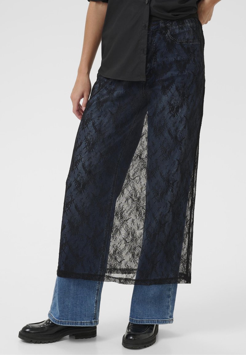 Jupe longue en dentelle noire avec une superposition transparente, un motif texturé et des fentes sur les côtés, portée par-dessus un pantalon en jean bleu. Bottes noires brillantes visibles.