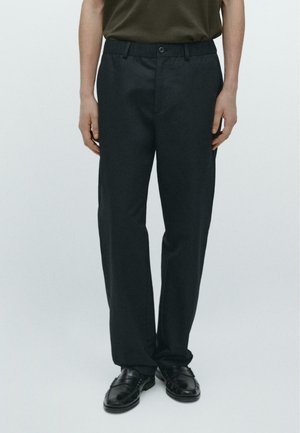 Mann trägt schwarze gerade geschnittene Hose, schwarze Lederschuhe und ein dunkelolivgrünes, in die Hose gestecktes Hemd, steht vor einem schlichten Hintergrund.