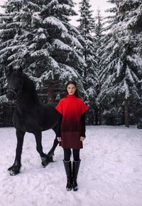Mujer con vestido de suéter rojo y botas negras, de pie en un bosque nevado junto a un gran caballo negro con nieve en sus patas.